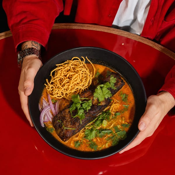 Hands holding a black bowl of Khao Soi with crispy noodles, braised short rib, and fresh herbs.