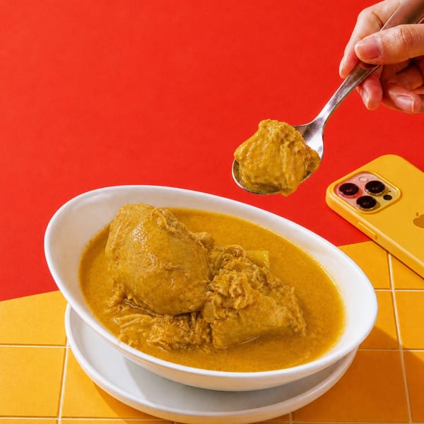 A hand spoons yellow chicken curry from a white bowl on a red and yellow tiled surface.