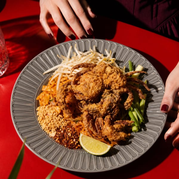 Hands reaching for a plate of crispy soft-shell crab pad Thai with bean sprouts, peanuts, and lime.