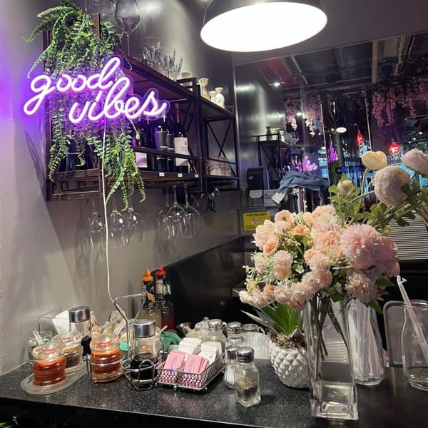 A purple neon "good vibes" sign hangs above a counter with condiments and flowers at Pink Pepper.