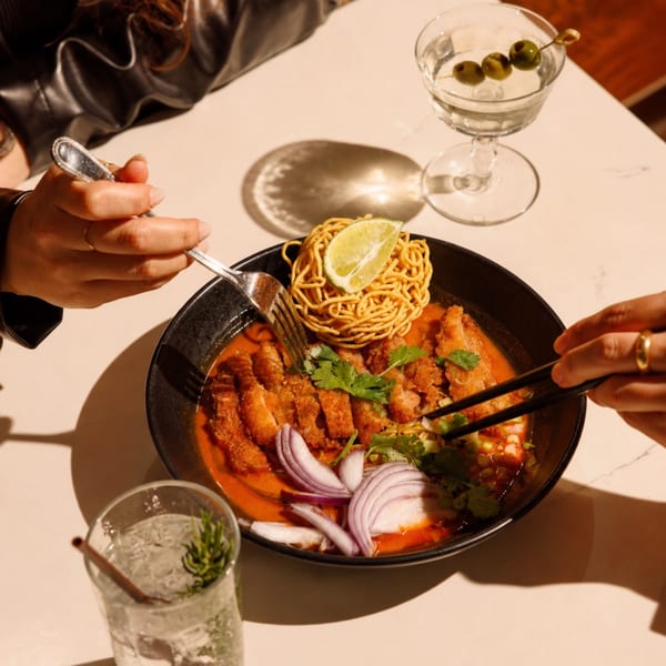 Two people sharing a bowl of red curry with crispy chicken, noodles, and lime, with cocktails.