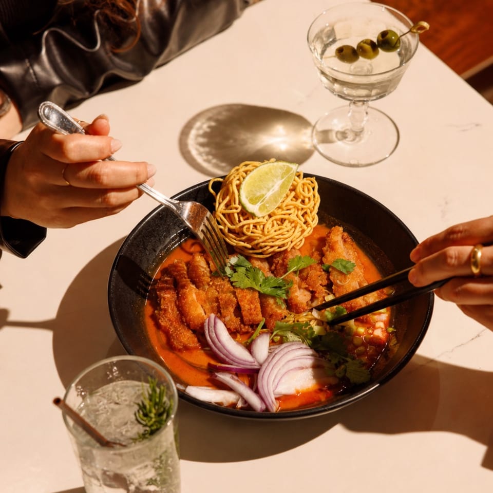 Two people sharing a bowl of red curry with crispy chicken, noodles, and lime, with cocktails.