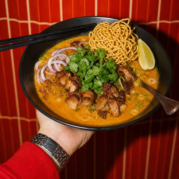 Hand holding a bowl of Khao Soi with chicken, crispy noodles, cilantro, red onion, and lime.