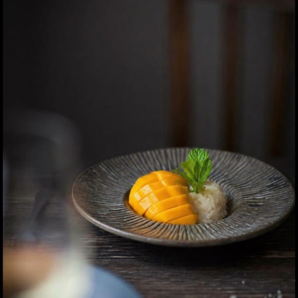 Mango sticky rice dessert with mint on a textured dark plate, on a wooden table.