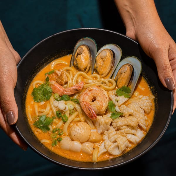 Hands holding a black bowl of seafood ramen with mussels, shrimp, squid, and scallops.
