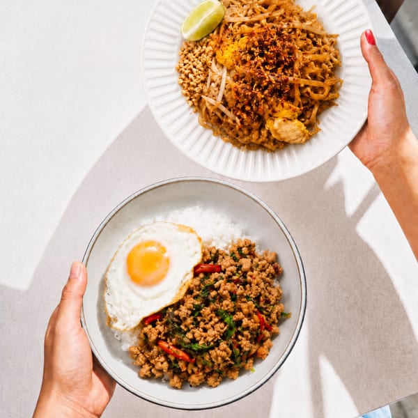 Hands holding two plates of Thai food: Pad Thai with lime, and rice with minced meat and a fried egg.