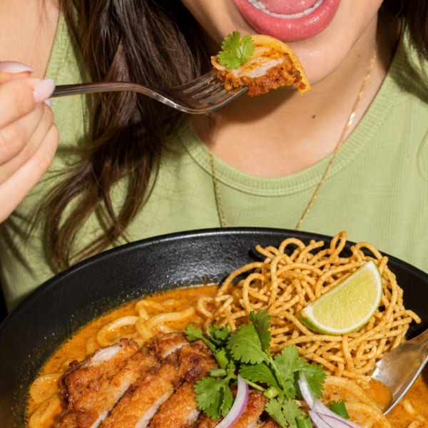 A person in a green shirt eats a spoonful of Khao Soi with chicken, noodles, and lime.