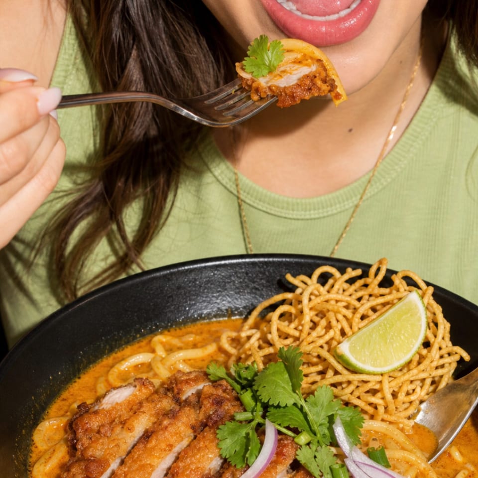 A person in a green shirt eats a spoonful of Khao Soi with chicken, noodles, and lime.