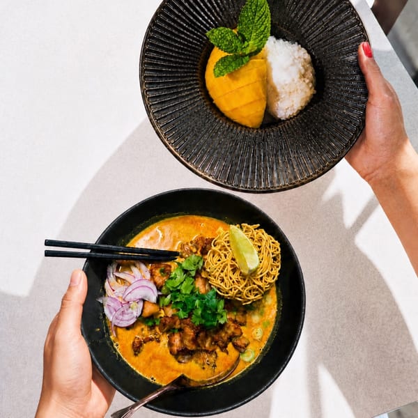 Hands holding a bowl of curry noodles and a plate of mango sticky rice.