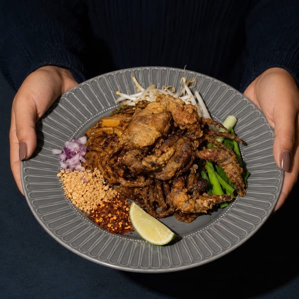 Hands holding a plate of Pad See Ew with crispy soft-shell crab, bean sprouts, and lime.