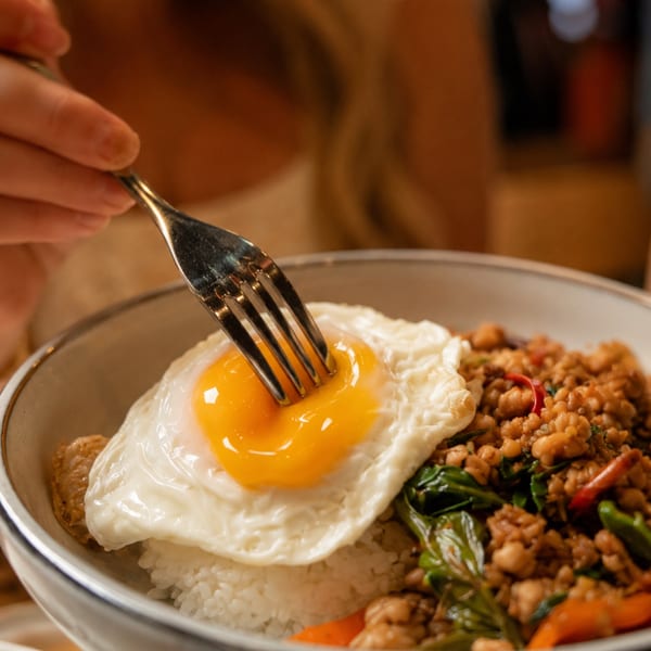 A fork breaks the yolk of a fried egg on a bowl of rice with minced meat and basil.