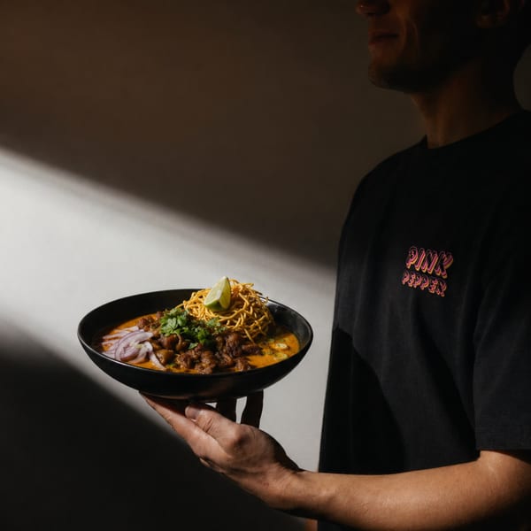 A person in a black Pink Pepper t-shirt holds a bowl of curry with noodles, lime, and red onion.