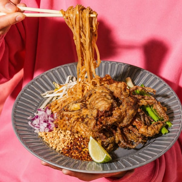 Chopsticks lifting noodles from a plate of Pad Thai with fried soft-shell crab, lime, and peanuts.